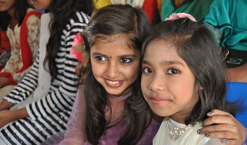Two young girls embrace at a Christmas event in Bangladesh, while wearing their best dresses with makeup on their faces.