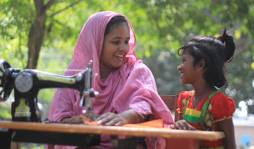A smiling woman in a pink scarf sits at a sewing machine outside, next to a smiling little girl in a red dress.