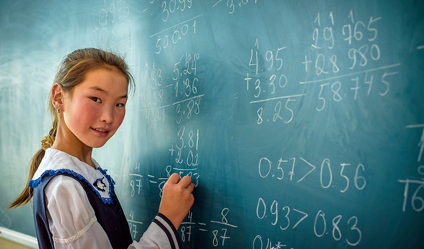 A girl looks toward the camera as she writes an addition problem on the green chalkboard