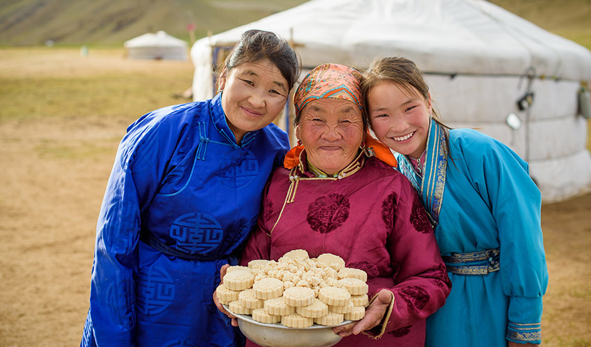 Three smiling women of different ages stand together, the oldest holding a plate of white circular patties
