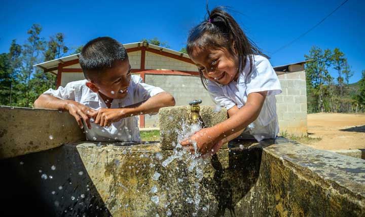A young girl in Honduras wearing school uniform splashes clean water as she washes her hands. She’s smiling broadly as her friend looks on.