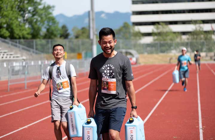 Men in shorts and t-shirts run on a sports track while carrying heavy blue water jugs.