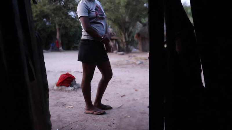 A teenage girl stands and waits outside her hut. She is wearing a white t-shirt and black skirt. Her face is not visible in the photo.