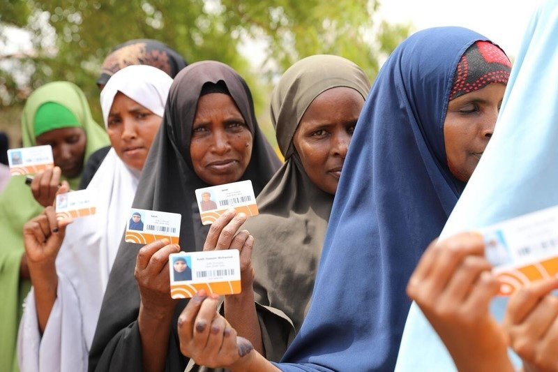 Women in Ethiopia wearing bright headscarves stand in line for cash assistance. They are holding up identification cards.