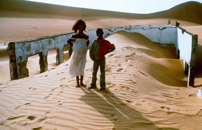 A boy and a pregnant woman stand in the desert, the walls of a derelict building around them.