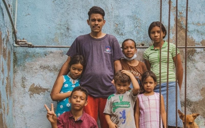 A Venezuelan refugee family stands together against  brick wall with peeling paint.