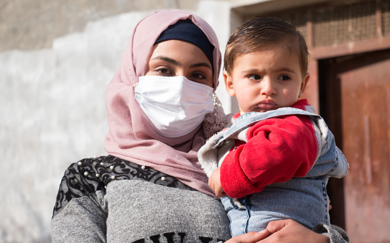 A woman wearing a medical face mask holds a toddler in her arms.