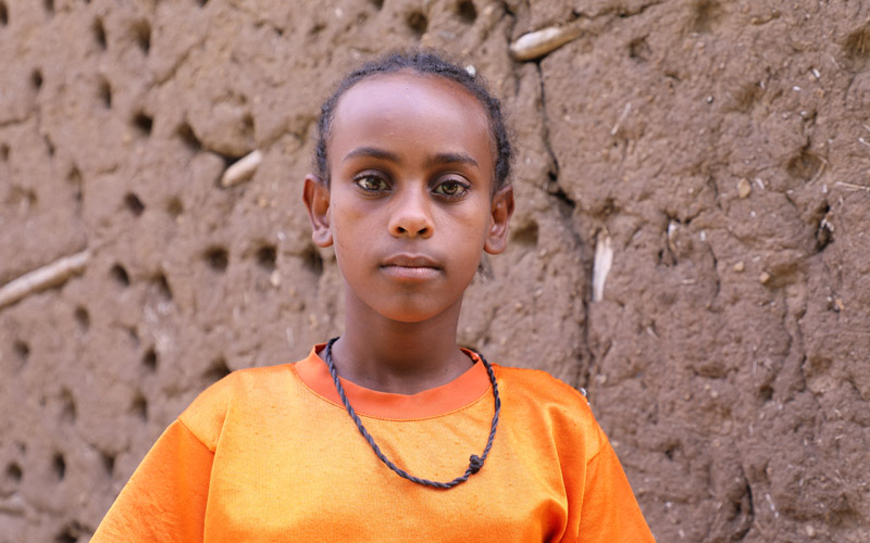 a young Ethiopian girl stands in front of a mud wall
