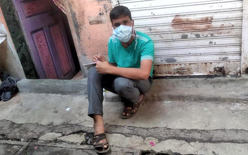 A teenage boy wearing a mask sits on the ground and writes on a notepad.