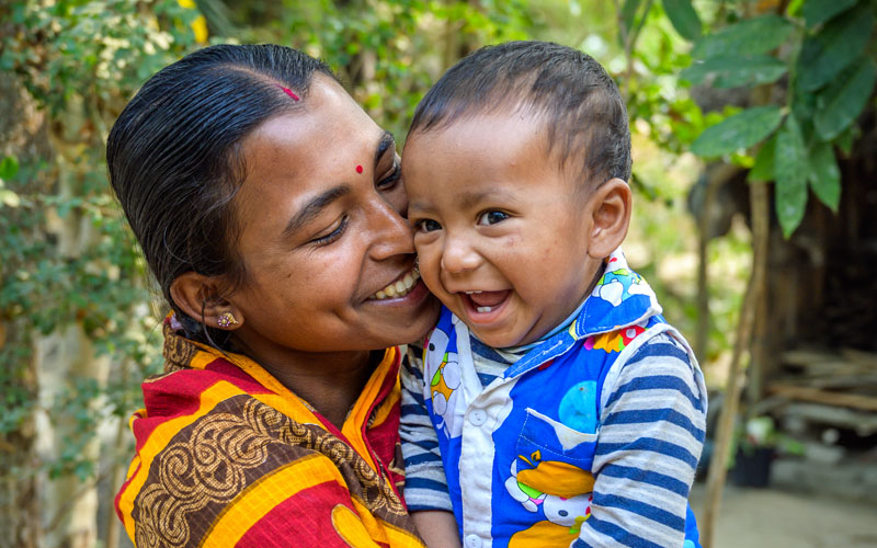 A Bangladeshi woman cuddles a baby close to her face. They are both smiling and laughing.