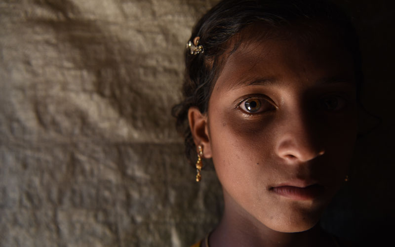 A close up image of a young girl from Myanmar. Half of her face is in shadow and she looks directly at the camera