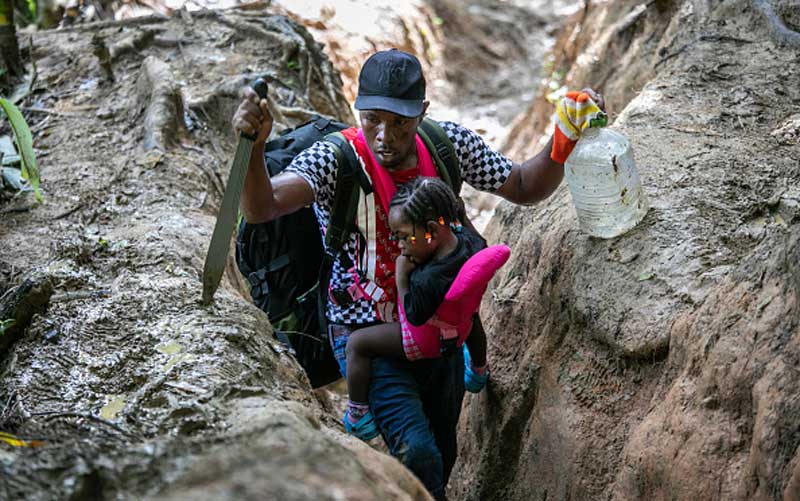 A man gasps for breath while trudging up a deep, muddy trench. He is carrying a little girl in a sling, as well as a machete and a water bottle.