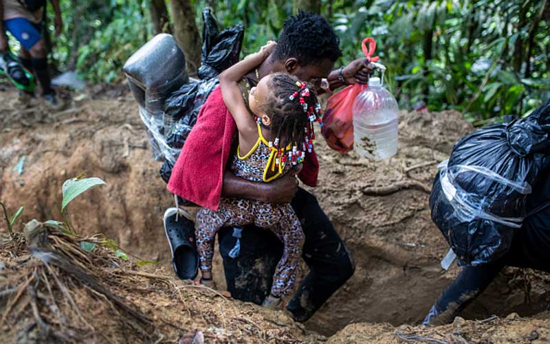 A man carries a preschool-aged girl with one arm, water in the other, and supplies on his back. He is walking through a deep, muddy trench in the jungle.