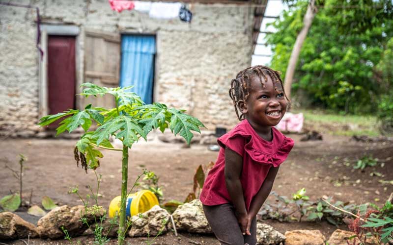 In Haiti, a small child smiles while standing beside some plants in her family’s garden.