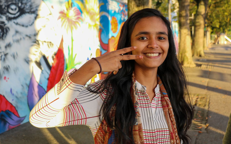 A young Bangladeshi girl poses for the camera near a colourful wall. She is smiling and giving the peace sign