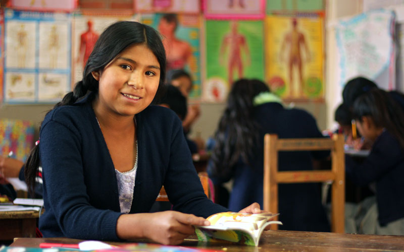 A young Bolivian girl sits at a desk with a book. She wears a navy blue sweater and smiles at the camera