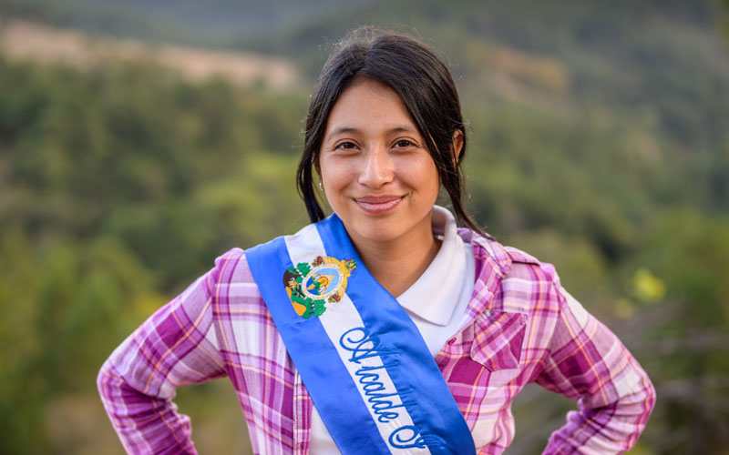 A young Honduran girl stands with hands on hips, smile on face. She wears a pink plaid shirt and a blue and white sash.