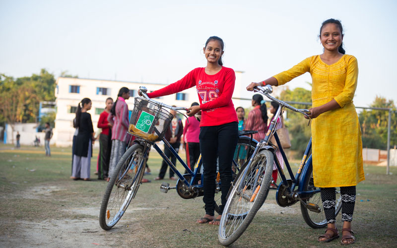 Two Indian girls stand next to bicycles. There is a group of girls standing behind them and they are near a school.