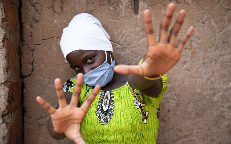 A young girl from Uganda wears a mask and holds her hands forward as if to say "Keep back."