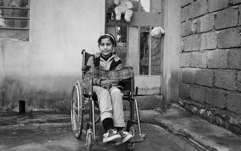 A young girl from Iraq sits in a wheelchair in a walled concrete courtyard.