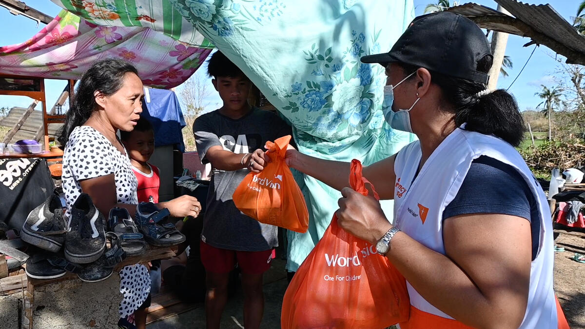 A woman in a World Vision vest hands bags of supplies to a family sheltering under a tarpaulin.