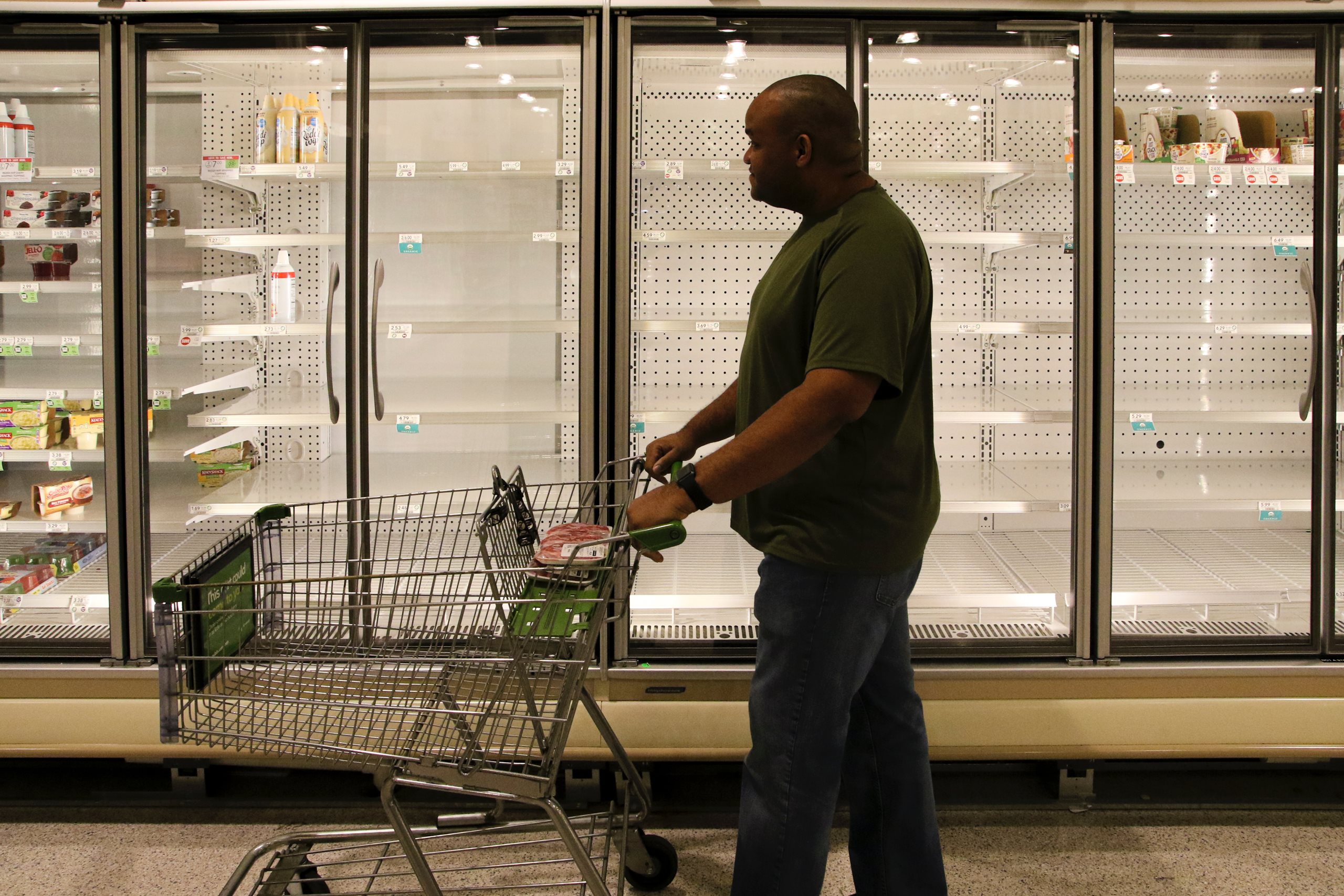 In North America, a man walks with his empty shopping cart past an empty grocery store fridge.