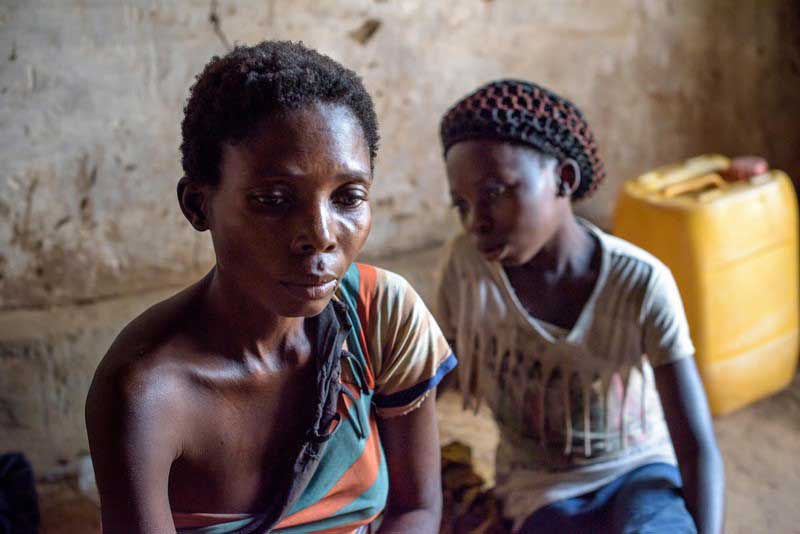 A mother and daughter sit quietly together, looking weary and despondent. The daughter is just an adolescent. The mother is wearing a torn shirt.