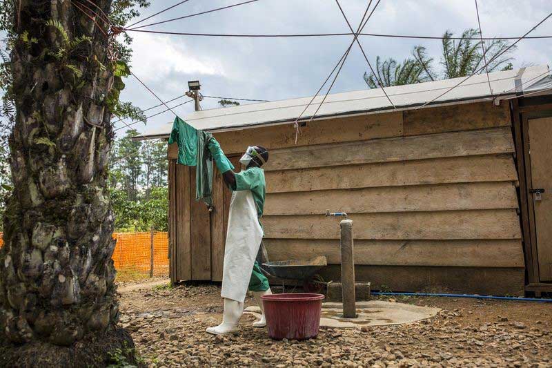 A health worker in plastic apron, gloves, mask and boots hangs sterilized materials on a line to dry outside a wooden hut.