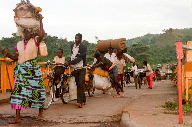 A surge of Rwandan people carrying large bundles and mattresses walk across a bridge in the same direction. They look rushed and serious.