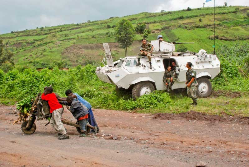 Peacekeepers rest by their military vehicle at the side of the road. On the road, several children struggle to push a wooden cart holding bunches of bananas.