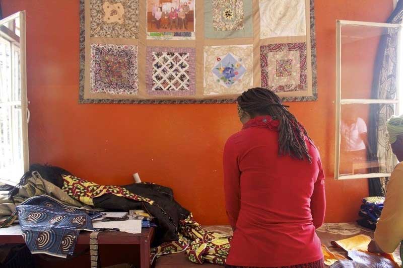 Women in the Democratic Republic of Congo have their backs to the camera, as they lean over a table, cutting and pinning colourful bits of fabric. A bright, hand-crafted quilt hangs on the wall behind them.