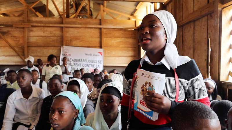 A young woman stands up in a crowded meeting, holding pamphlets about Ebola. She addresses the group. Her posture and face look confident.