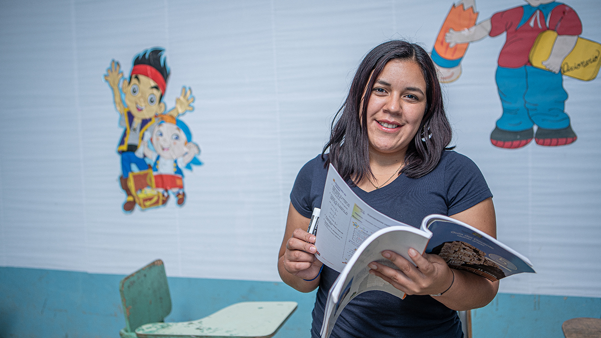 A woman standing in a classroom, turning the page of a book as she smiles at the camera.