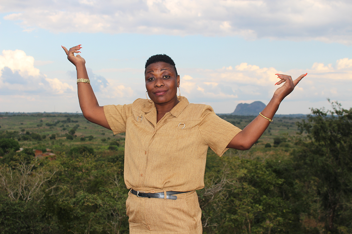 A woman wearing a light brown uniform stands in front of a landscape with her arms in the air in a gesture of pride and accomplishment.