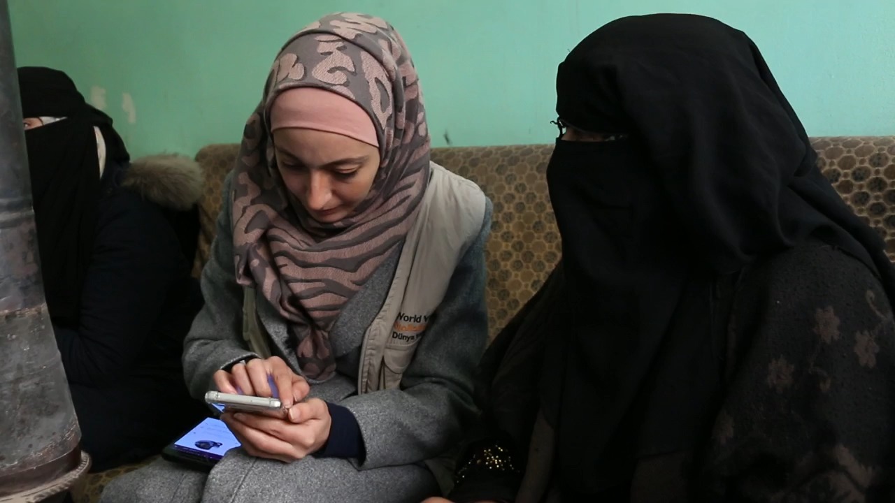 A staff member wearing a hijab sits on a couch with two women in niqabs, talking with them and looking at her phone.