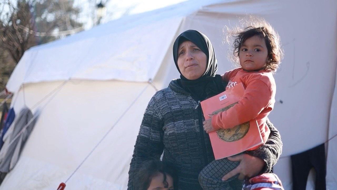 A woman stands outside a tent, carrying her young child who is holding a book.