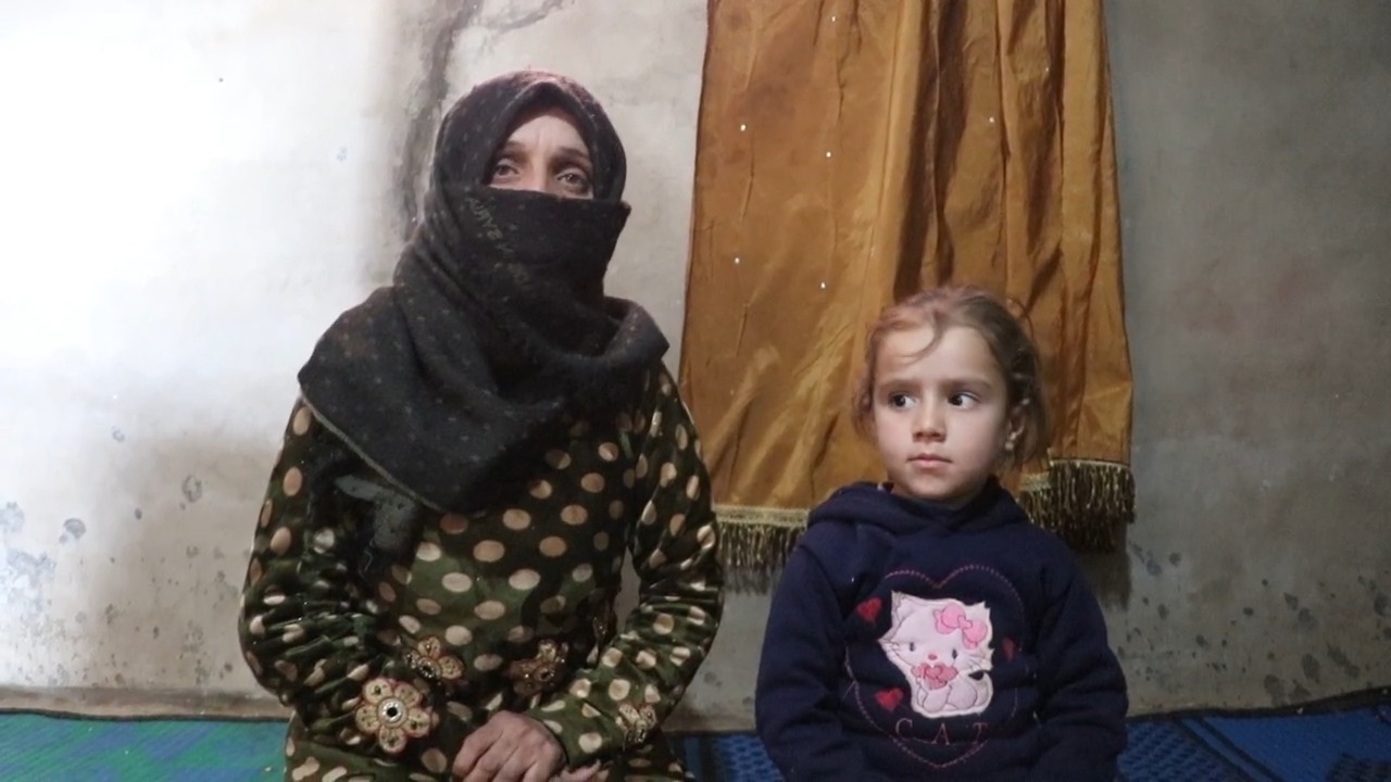 Abdo sits with her young daughter beside her, and she is wearing a gray hijab and green and beige polka dot dress.