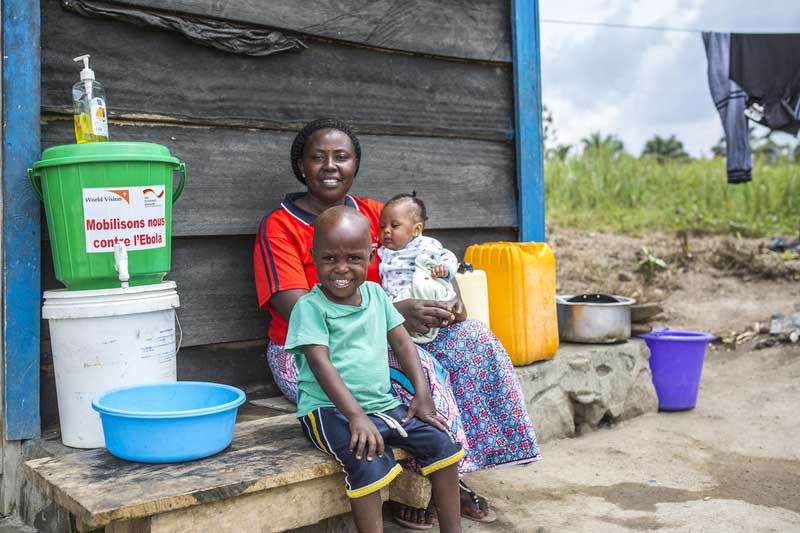 A woman holding a baby and a small boy sit in front of a wooden home with water buckets next to them.