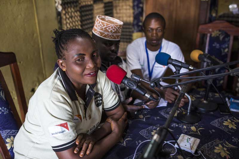 A woman and two men from the DRC sit in a radio studio recording a radio show.