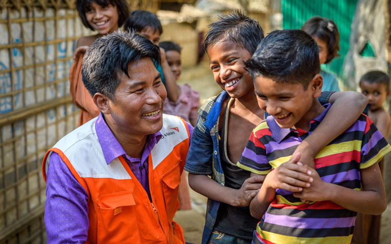 A man in an orange and white World Vision vest smiles at two boys.