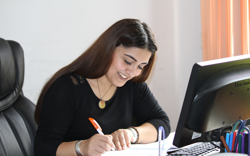 A woman in professional attire sits at a desk and writes, smiling.