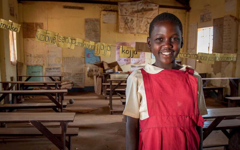 A young girl wearing a red school uniform dress stands in front of her classroom smiling.