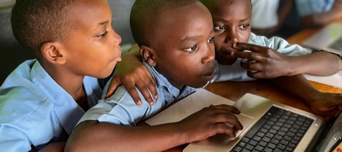 Three inquisitive young boys in blue school uniforms crowd around a laptop computer.