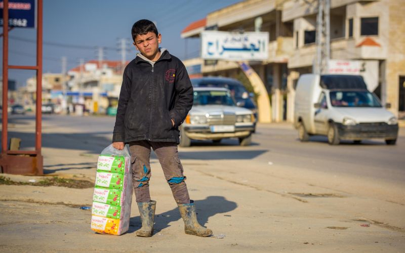 An adolescent boy stands on the street holding a large multi-pack of tissue boxes.