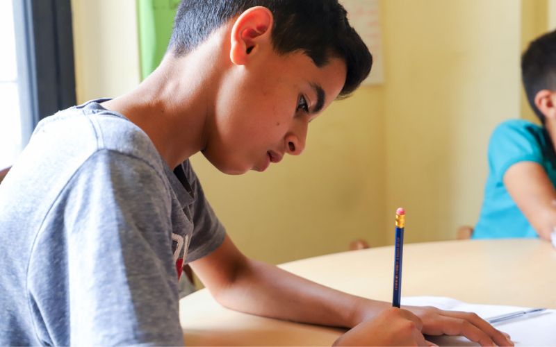 An adolescent boy sits at a desk, writing in a notebook with a pencil.