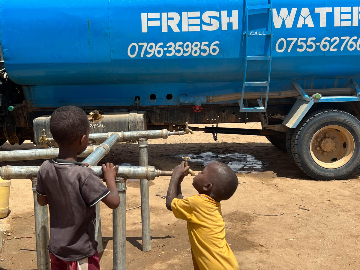 Two children wait by a water tap, looking toward a large water truck in the background.