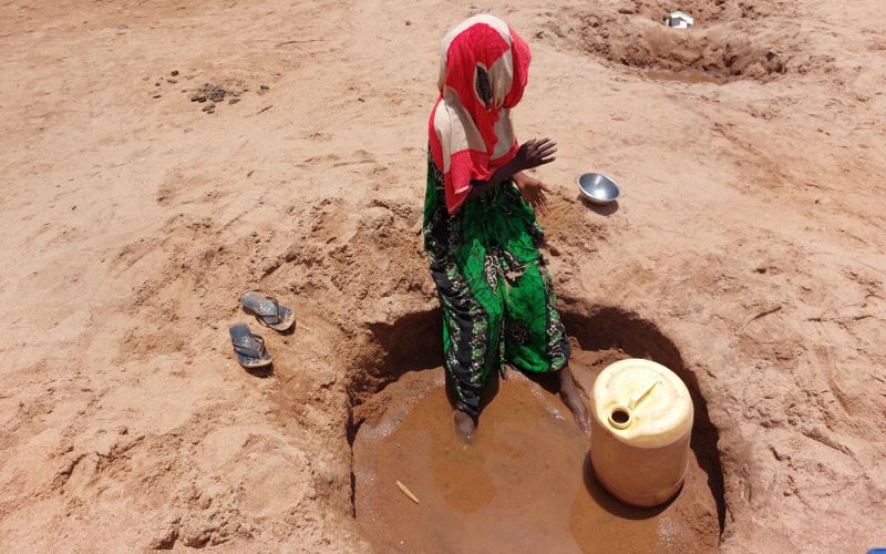 In Somalia, a woman sits on the edge of a hole dug in a dry river bed. Her feet sink into the mud at the bottom.