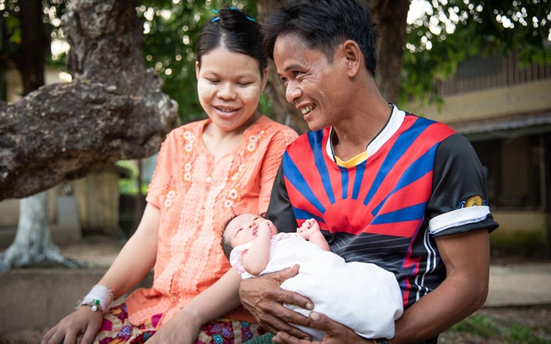 A man from Myanmar smiles holding a baby, and a woman sits next to him smiling down at the baby.