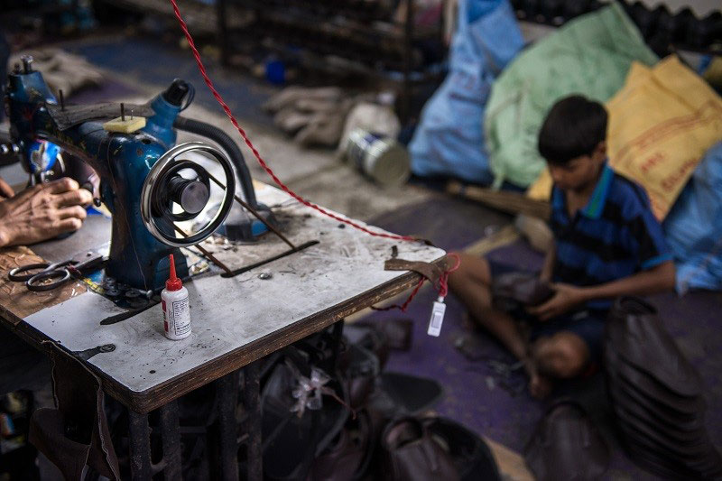 A sewing machine in the foreground looms large. A small boy sits cross-legged on the floor behind, bent over his sewing.