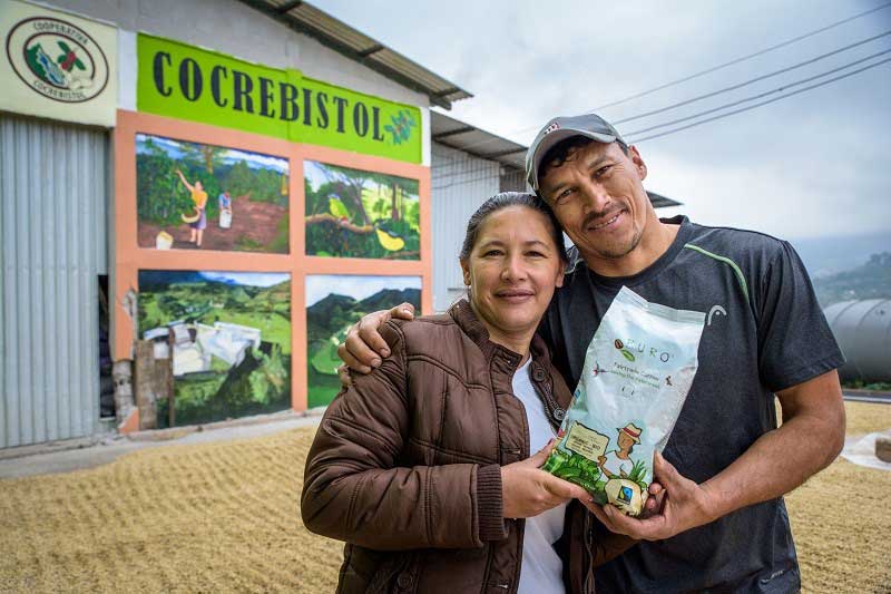 A man and woman in Honduras proudly hold a bag of coffee beans. They are standing in front of their business.